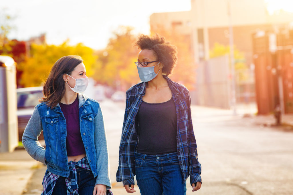 Two female friends walking down a Brooklyn alley wearing face masks on a sunny Autumn day, having a quiet conversation.