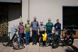 Group of six pople standing in front of a building, two pose with bikes