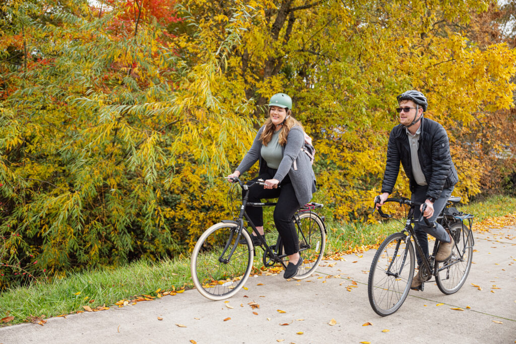 Two people biking together. The trees are turning for fall and both cyclists wear clothes for an office job.